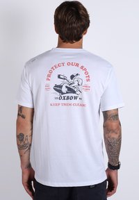 T-shirt en coton blanc avec un graphique au dos comportant un texte rouge et noir. Le design inclut une illustration d'un personnage avec une planche de surf.