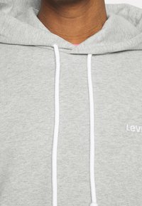 Felpa grigia con cappuccio, dotata di laccetti bianchi e logo "Levi's" ricamato. Tessuto morbido e texture. Vista ravvicinata.