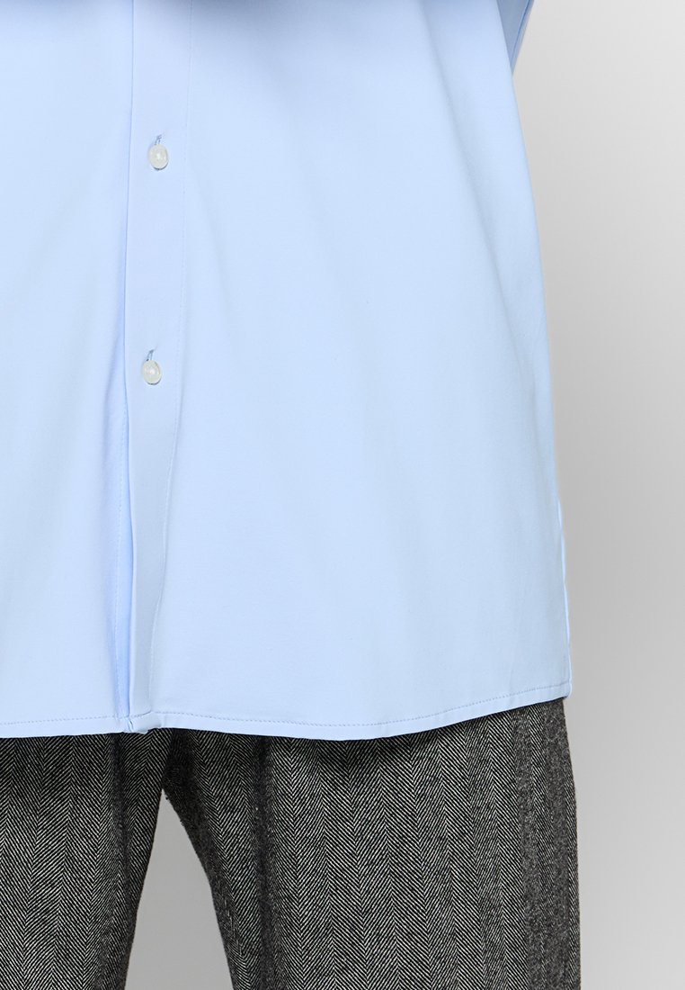 Chemise bleu clair à manches longues en tissu lisse, avec un col classique et un ourlet arrondi, associée à un pantalon sombre à motif chevrons.