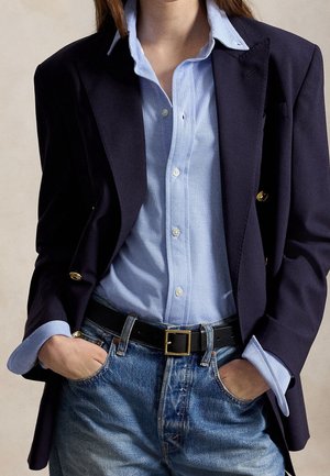 Personne portant un blazer bleu marine, une chemise bleu clair boutonnée, un jean bleu et une ceinture noire, les mains dans les poches.