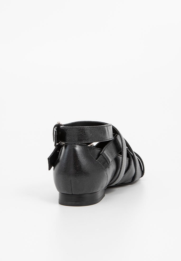 Sandalia de cuero negro con tacón plano, que presenta tiras cruzadas y una correa ajustable en el tobillo. Textura suave y diseño minimalista.