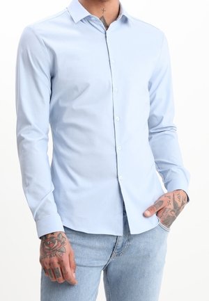 Camicia slim fit azzurro chiaro con bottoni, indossata senza essere infilata nei pantaloni, abbinata a jeans chiari, con mani e collo tatuati in vista.