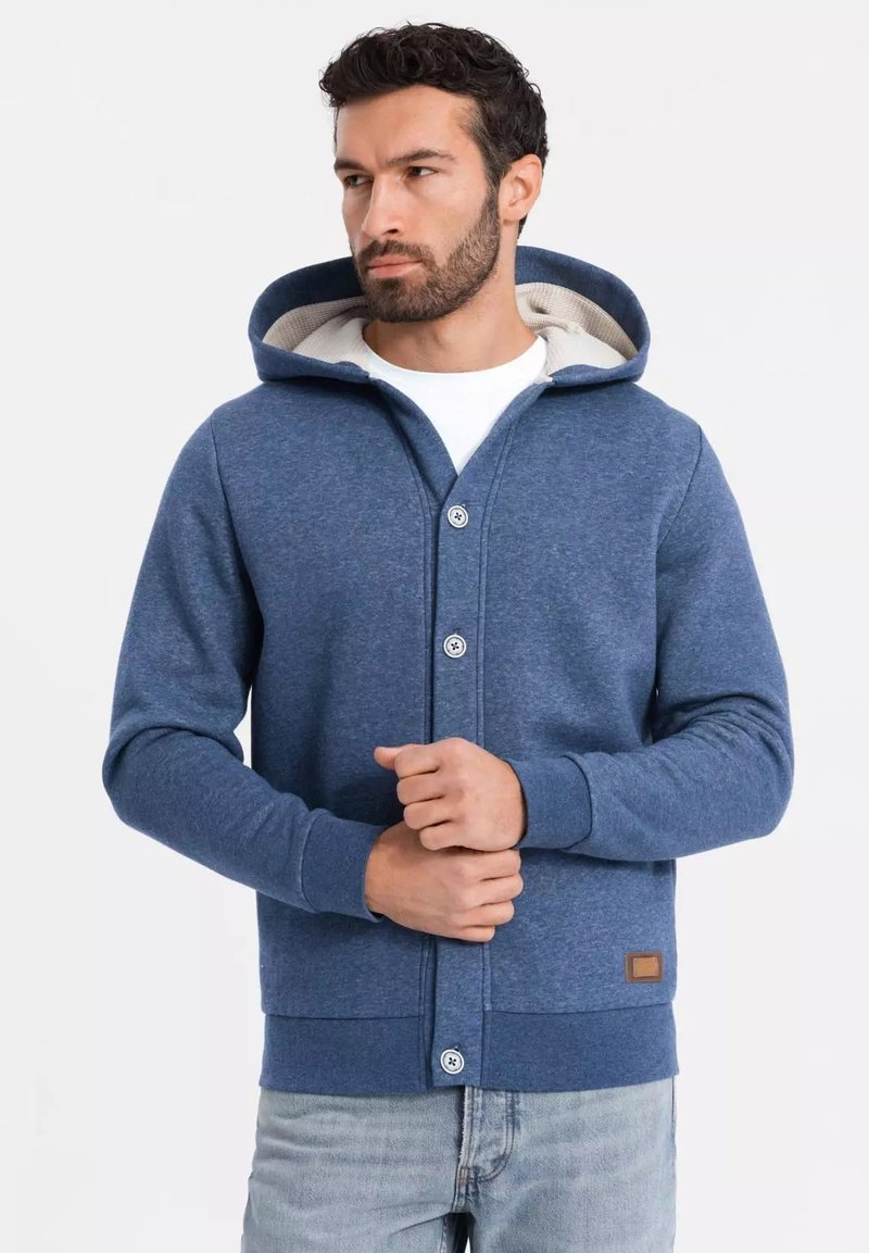 Sudadera con capucha azul marino con cierre de botones, fabricada en un material suave, con forro interior crema y una pequeña etiqueta de cuero marrón en el dobladillo.