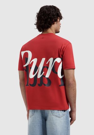 Man met krullend donker haar, draagt een rood T-shirt met korte mouwen met grote witte en zwarte tekst "Pure" op de achterkant en lichtblauwe spijkerbroek.