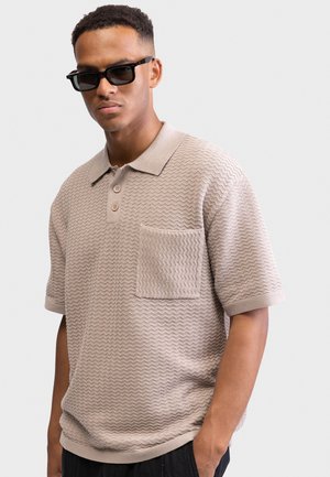 Άνδρας που φοράει υφασμάτινο μπεζ κοντομάνικο πόλο με μπροστινή τσέπη και μαύρα γυαλιά ηλίου, στέκεται μπροστά σε απλό ανοιχτό φόντο.