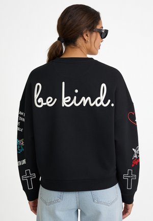 Frau mit Sonnenbrille und schwarzem Sweatshirt mit "be kind." in großen weißen Buchstaben auf dem Rücken, bunte Aufnäher an den Ärmeln, helle Jeans.