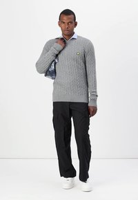 Suéter gris de punto trenzado con puños acanalados, combinado con pantalones cargo negros y zapatillas blancas. Fondo claro, accesorios mínimos.