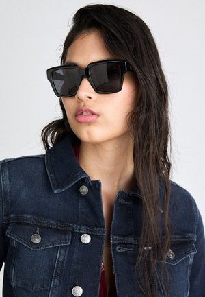 Occhiali da sole neri con montatura spessa, lenti scure e sottili dettagli in oro, abbinati a una giacca di jeans scura con bottoni argentati.