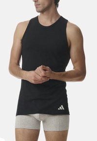 Schwarzes Tanktop aus weichem Stoff, mit einem runden Halsausschnitt und einem weißen Logo unten links. Getragen mit hellgrauen Boxershorts.