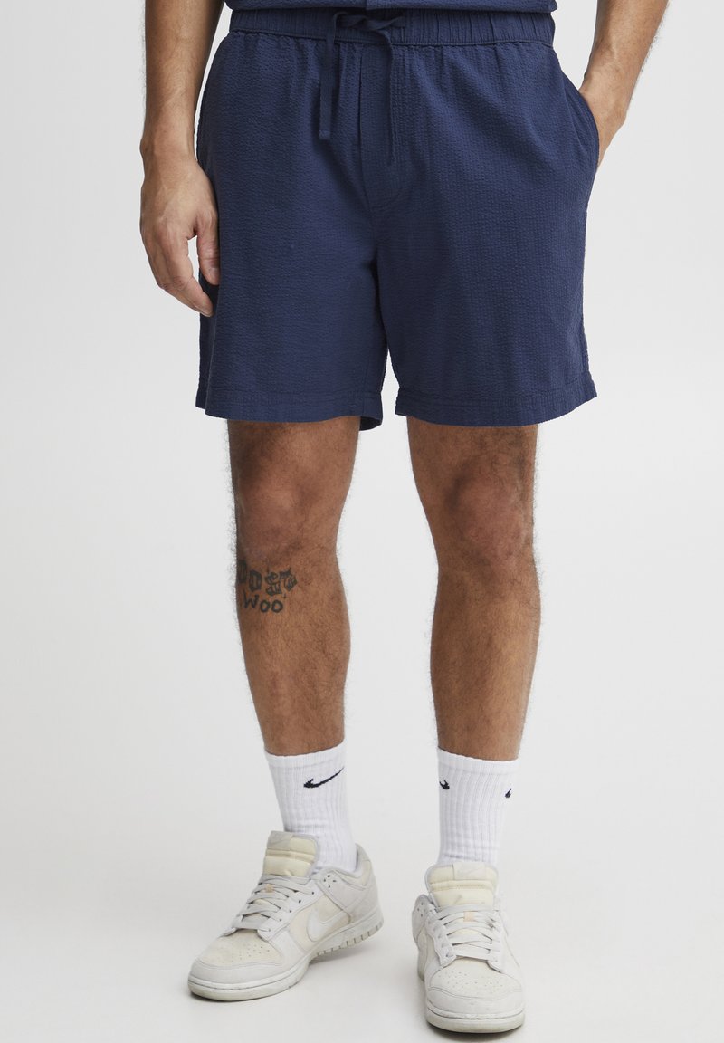 Marineblaue Shorts aus strukturiertem Stoff, mit elastischem Bund, Kordelzug und seitlichen Taschen. Tragen mit weißen Nike-Socken und Sneakers.
