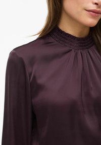 Satinlilla bluse med høj smocket halsudskærning, lange ærmer og subtile plisseringer. Glat tekstur og elegant glans hele vejen igennem.