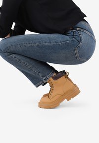 Botas de tobillo de cuero marrón claro con diseño de cordones, suela de goma resistente y ribete de tobillo en marrón oscuro. Combinadas con jeans de mezclilla azul.