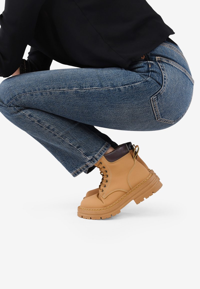 Botas de tobillo de cuero marrón claro con diseño de cordones, suela de goma resistente y ribete de tobillo en marrón oscuro. Combinadas con jeans de mezclilla azul.