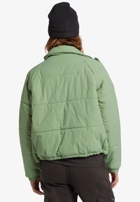 Giacca puffer verde chiaro con una vestibilità rilassata, caratterizzata da cuciture orizzontali, un colletto e orlo elasticizzato. Indossata con un berretto nero.