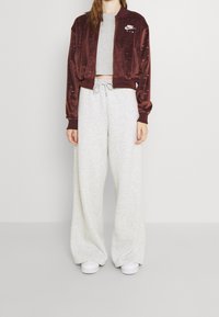 Burgundsko červená sametová bomber bunda s logem Nike, spárovaná s volnými šedými tepláky, které mají elastický pas a stažené kotníky.