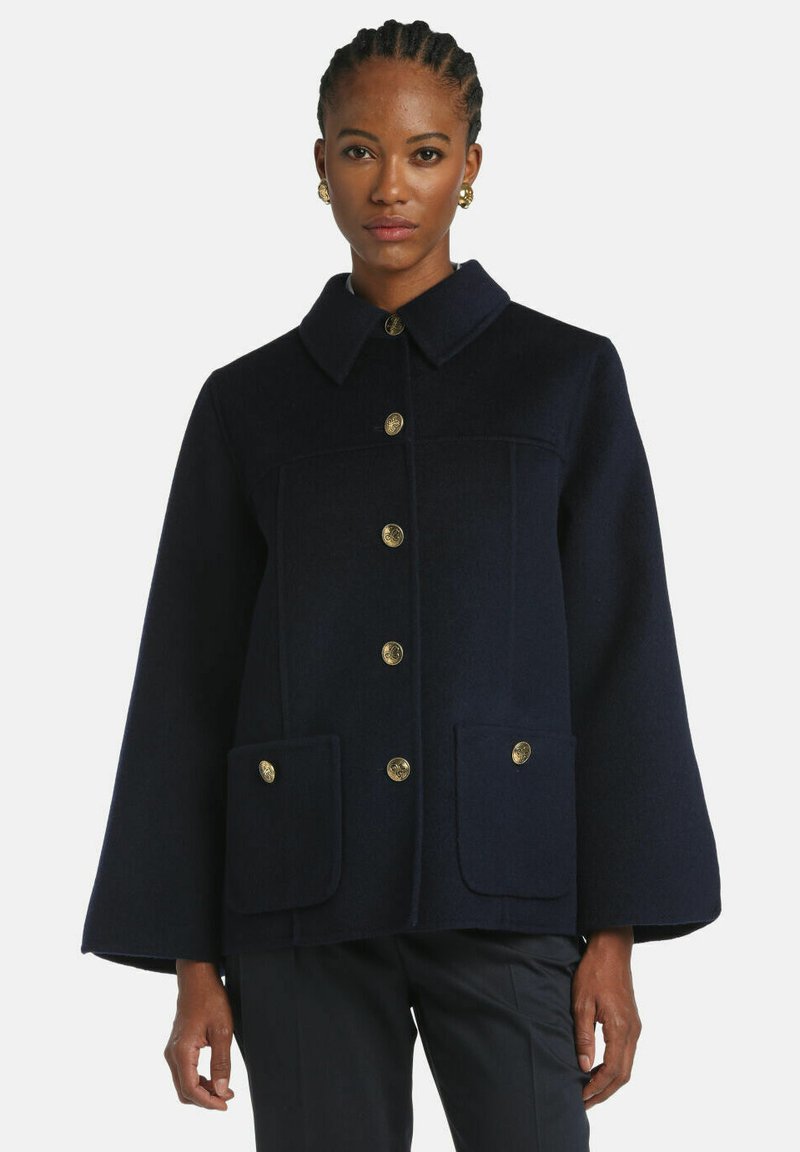 Námornícky modrý vlnený kabát s bodovým golierom, zlatými gombíkmi, dvoma prednými vreckami a širokými rukávmi. Hladká textúra, štruktúrovaný dizajn.