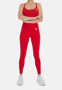 Soutien-gorge de sport rouge et leggings taille haute assortis en tissu extensible. Accents de logo blanc sur la poitrine et la cuisse, portée avec des baskets blanches.