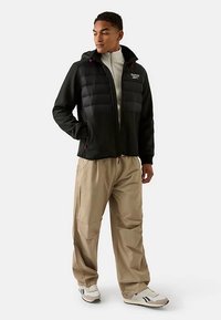 Schwarze Reißverschlussjacke mit gepolstertem Vorderteil, verstellbarer Kapuze und roten Akzenten, kombiniert mit locker sitzenden beigen Hosen und weißen Turnschuhen.