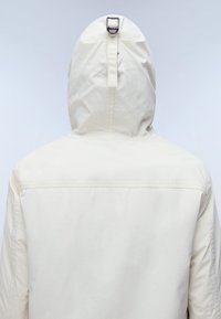 Vista trasera de una persona que lleva una chaqueta con capucha de color crema con un detalle de hebilla metálica en el centro trasero de la capucha.