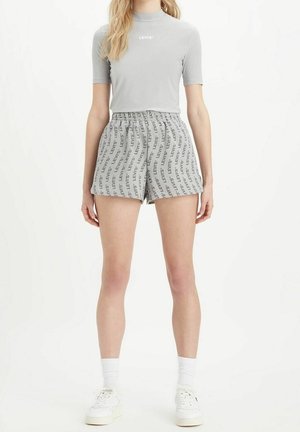 Kvinde stående iført en lysegrå, tætsiddende Levi's kortærmet top og elastik i taljen shorts med Levi's logo-print, hvide sokker og hvide sneakers.