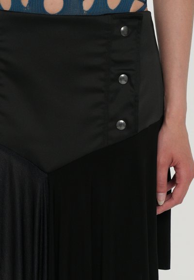 Jupe noire avec des textures satinées et plissées, présentant un design diagonal et trois boutons métalliques à la taille.