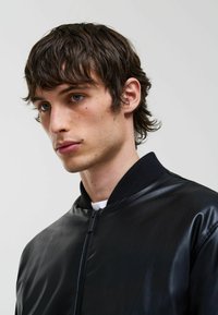 Jachetă bomber din piele neagră, cu guler ribbed și fermoar. Textură netedă, design ajustat, purtată peste o cămașă albă.