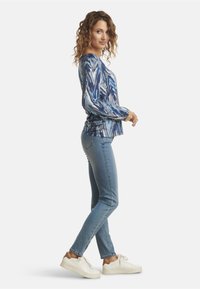 Blusa a maniche lunghe di colore blu a motivo con una texture seta, abbinata a jeans skinny azzurri e sneakers bianche. Vestibilità rilassata con dettagli arricciati.