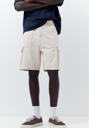 Bărbat purtând pantaloni scurți cargo bej, șosete albe, adidași maro și o cămașă albastru marin cu brațele încrucișate, stând în fața unui fundal simplu.