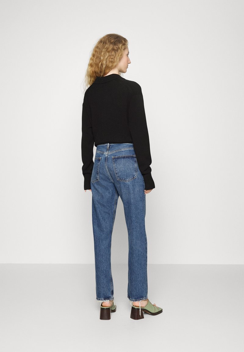 Un pull noir, un jean bleu taille haute avec une coupe droite, ourlet effiloché, associé à des mules à talons verts, vu de dos.