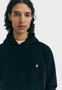 Čierna mikina s kapucňou, vybavená klokankovým vreckom a malým oranžovým grafickým motívom slnka na hrudi, vyrobená z mäkkej, textúrovanej látky s voľným strihom.