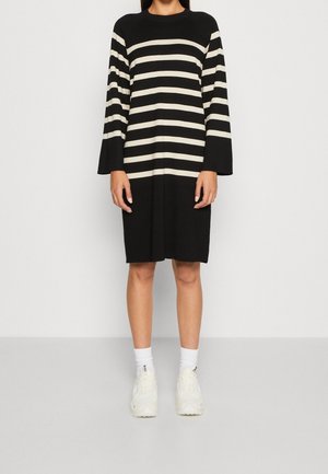 Kvinde iført en sort kjole med hvide vandrette striber på ærmer og bryst, hvide sokker og hvide sneakers, stående på en grå gulv.