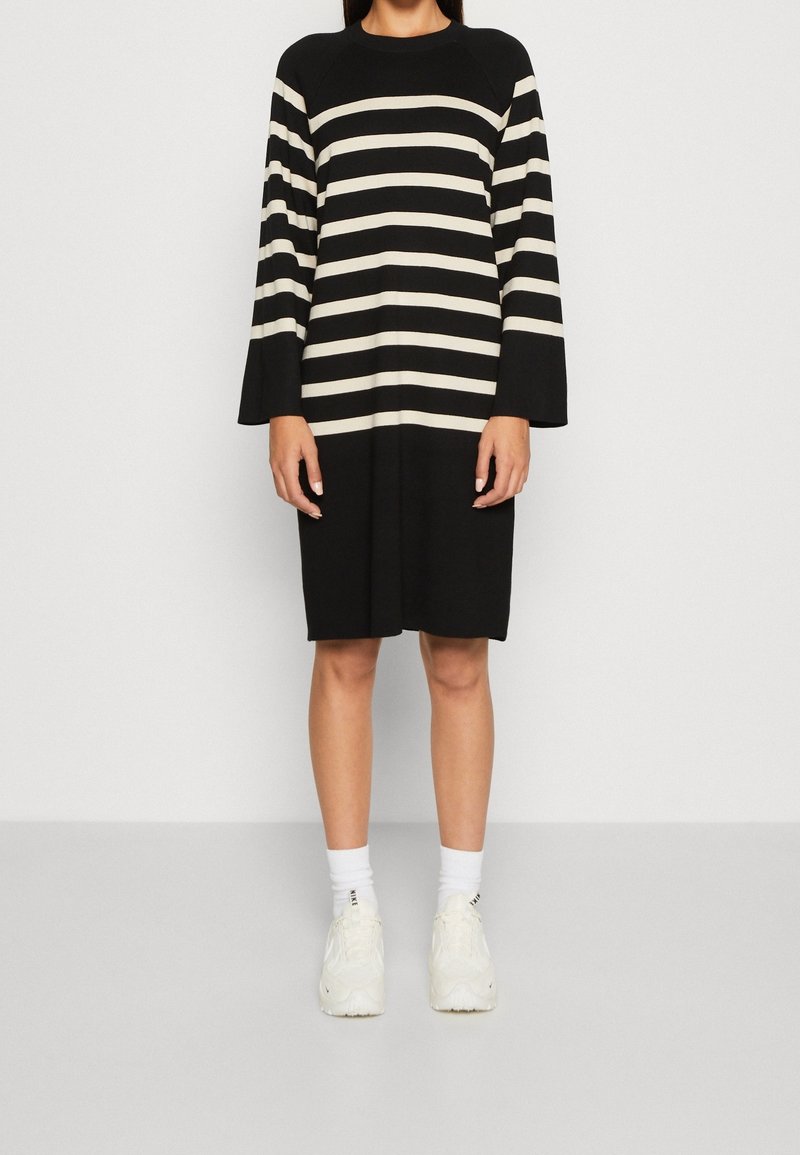Femme portant une robe noire avec des rayures horizontales blanches sur les manches et la poitrine, des chaussettes blanches et des baskets blanches, debout sur un sol gris.