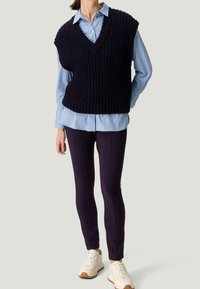 Marineblauer, gestrickter ärmelloser Pullover mit V-Ausschnitt, getragen über einem hellblauen, gestreiften Button-Up-Hemd, kombiniert mit dunklen Hosen und weißen Sneakers.