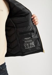 En person håller öppet en svart puffjacka och visar dess insida samt etiketter som beskriver materialvård och kvalitet.