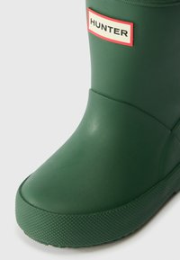 Bota de chuva de borracha verde com sola texturizada, bico arredondado e etiqueta de logo em branco e vermelho do lado lateral. Acabamento suave e mate.