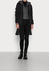 Abrigo negro de sarga sobre una chaqueta con capucha gris, pantalones ajustados negros y botas negras gruesas. Presenta puños contrastantes con patrones de rayas.
