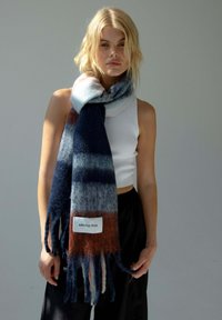 Écharpe en tricot avec des rayures bleues, grises et rouille, bordée de franges. Étiquette indiquant "ARCTIC FOX". Matière douce et texturée en laine.