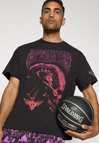 Sort grafisk t-shirt med et rosa kranie-design; korte ærmer, rund hals. Modellen holder en sort Spalding basketball med grå accenter.