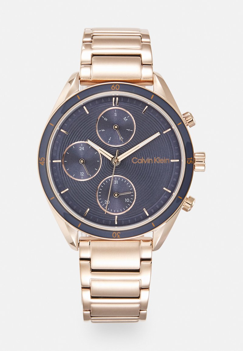 Montre Calvin Klein avec un cadran bleu foncé à motifs texturés, aiguilles dorées et sous-cadrans ; équipée d'un bracelet en métal doré.