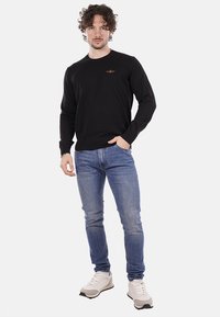 Maglione nero a maniche lunghe con scollo rotondo e logo sottile, abbinato a jeans blu e sneakers bianche con dettagli scuri.