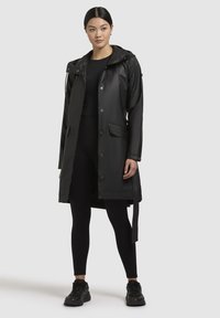 Zwarte waterdichte regenjas met capuchon, drukknoopjes, voorzakken en een strikdetail in de taille, gedragen over zwarte leggings en sneakers.
