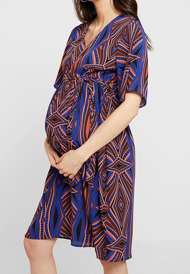 Femme enceinte portant une robe mi-longue avec des motifs géométriques bleus, marron et orange, tenant son ventre avec les deux mains.