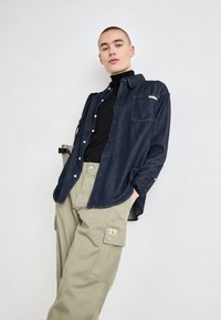 Camisa de mezclilla oscura con botones y un bolsillo en el pecho, llevada sobre un suéter de cuello alto negro, combinada con pantalones cargo beige claros que tienen bolsillos laterales.