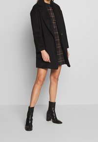 Svart kashmirblandad kappa med snibblagd krage, långa ärmar och öppen framsida. Kombinerad med en brun rutmönstrad klänning och knähöga blanka svarta stövlar.