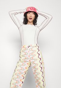 Top branco de manga longa com costura roxa, combinado com calças amarelas com padrões ondulados multicoloridos e um chapéu de balde vermelho com padrão.