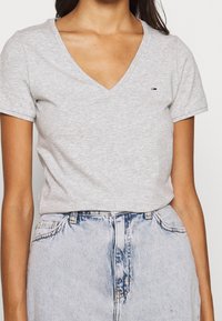 Grå kortärmad tröja med V-hals, tillverkad av mjuk bomull, med en liten logga på vänster bröst och en figursydd design.