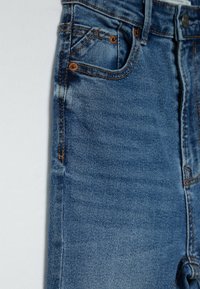 Gros plan d'un jean en denim bleu montrant la poche avant avec des rivets en cuivre, la ceinture, et une texture délavée sur le tissu.