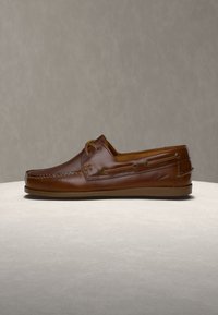 Καφέ δερμάτινο παπούτσι τύπου boat shoe με ραφές moc toe και λεπτομέρεια κορδονιών σε ουδέτερη επιφάνεια και φόντο.
