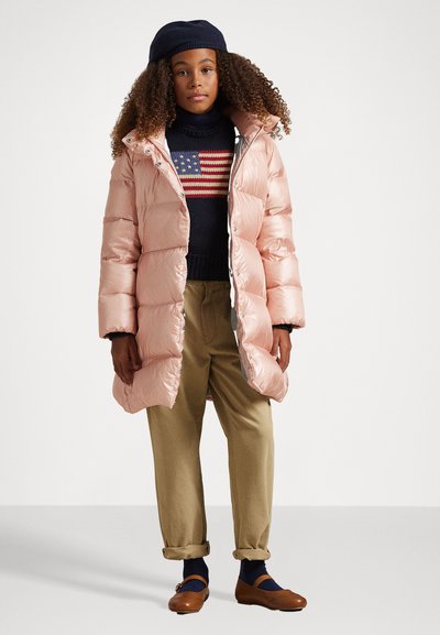 Casaco acolchoado rosa com capuz, grandes secções acolchoadas e peças em prata. Usado sobre um suéter azul-marinho com um padrão em vermelho e branco e calças bege.