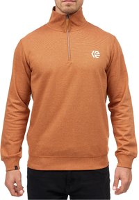Homme portant un pull à fermeture éclair quart couleur rouille avec un logo blanc sur la poitrine gauche et un pantalon noir, debout, les bras détendus.